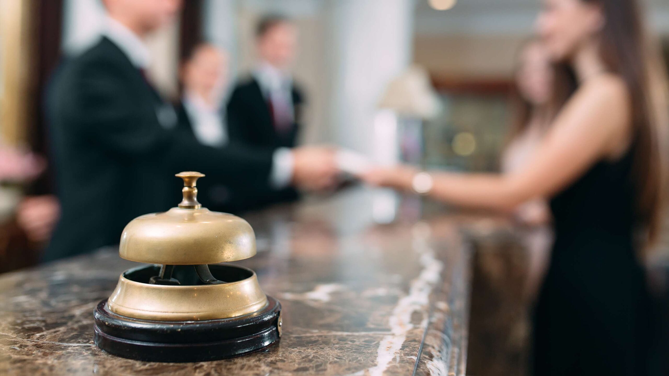 Primer plano de un timbre dorado de recepción sobre un mostrador de mármol en un hotel, con personal atendiendo a una huésped al fondo de manera desenfocada.