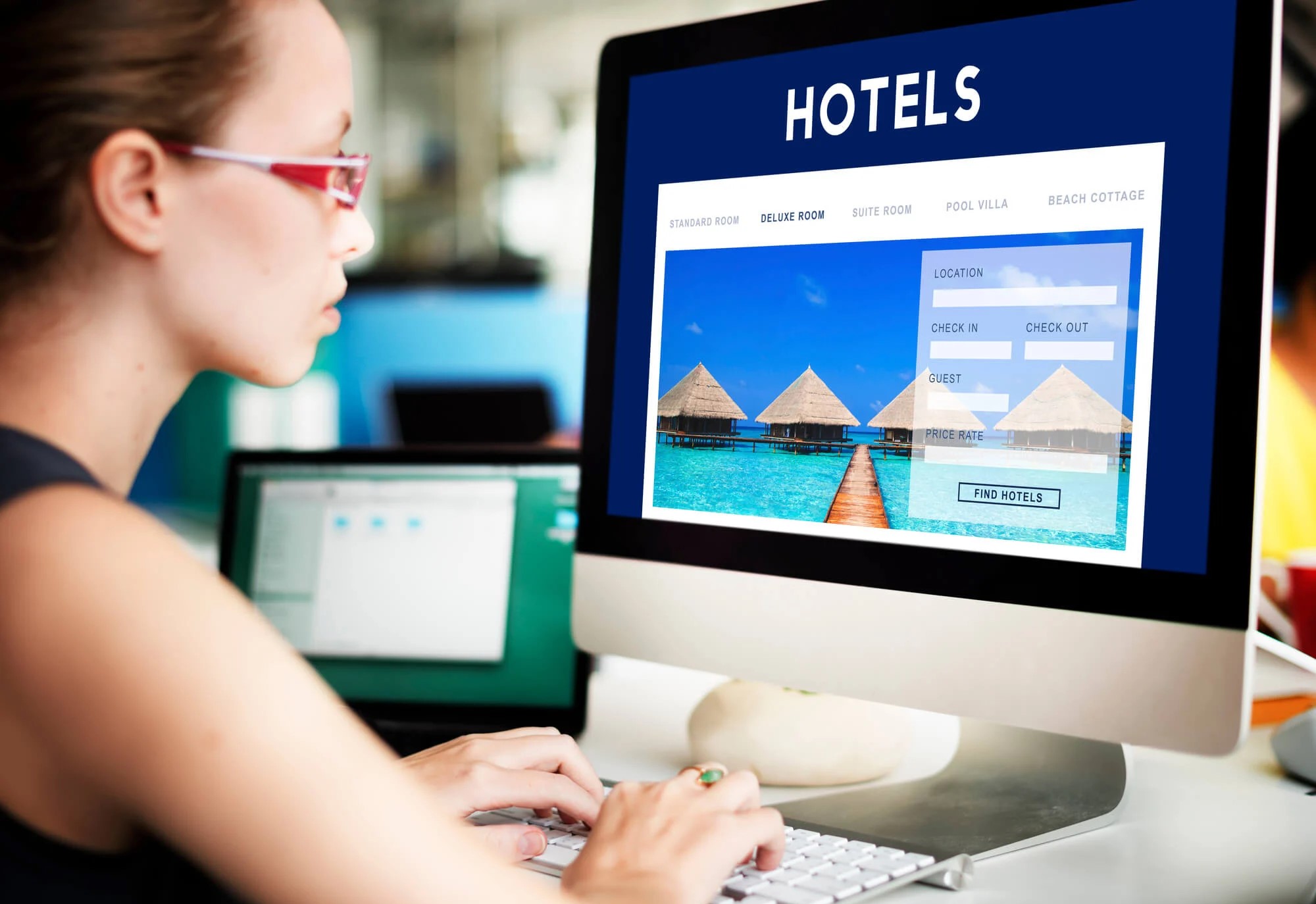 Mujer joven con gafas rojas utilizando una computadora de escritorio para buscar hoteles en línea; en la pantalla se muestra un portal de reservas con la imagen de bungalows sobre el agua en un destino tropical.